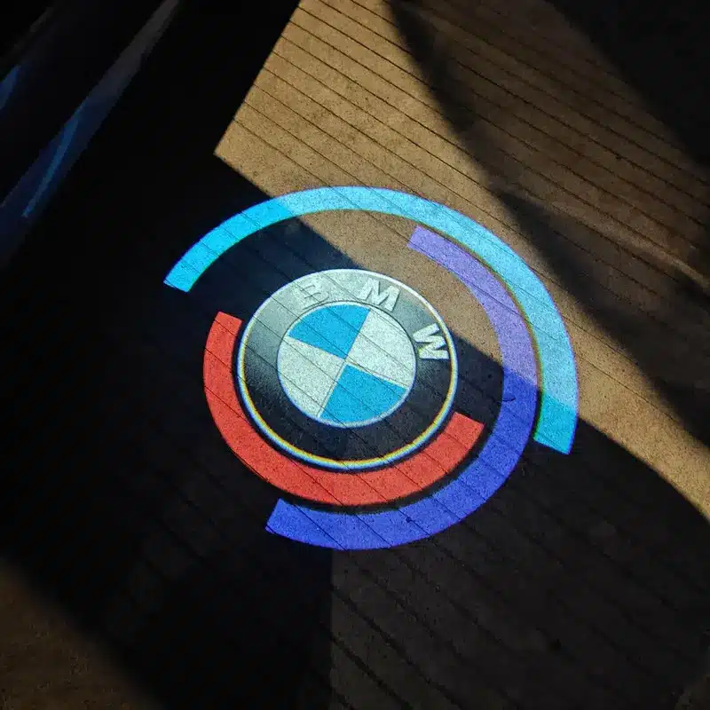 BMW 50th Anniversary Door Lights Projected BMW logo with blue and red curved lines on a dimly lit, textured surface, creating a modern and sleek visual effect.